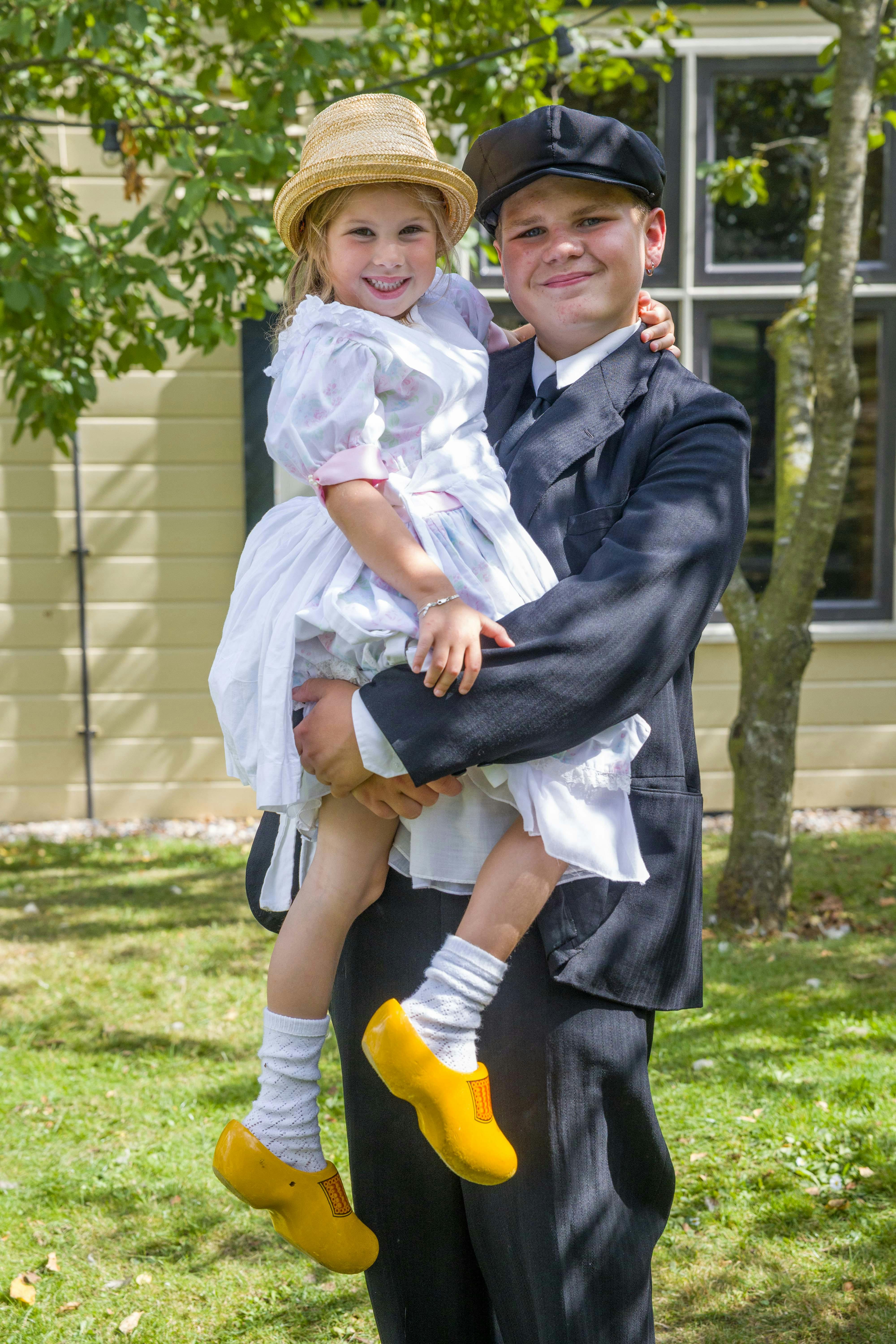 Ein Junge in einem dunklen Anzug und einer Mütze hält ein lächelndes Mädchen in einem hellen Kleid und einem Strohhut im Freien, in der Nähe eines Gebäudes und von Bäumen.