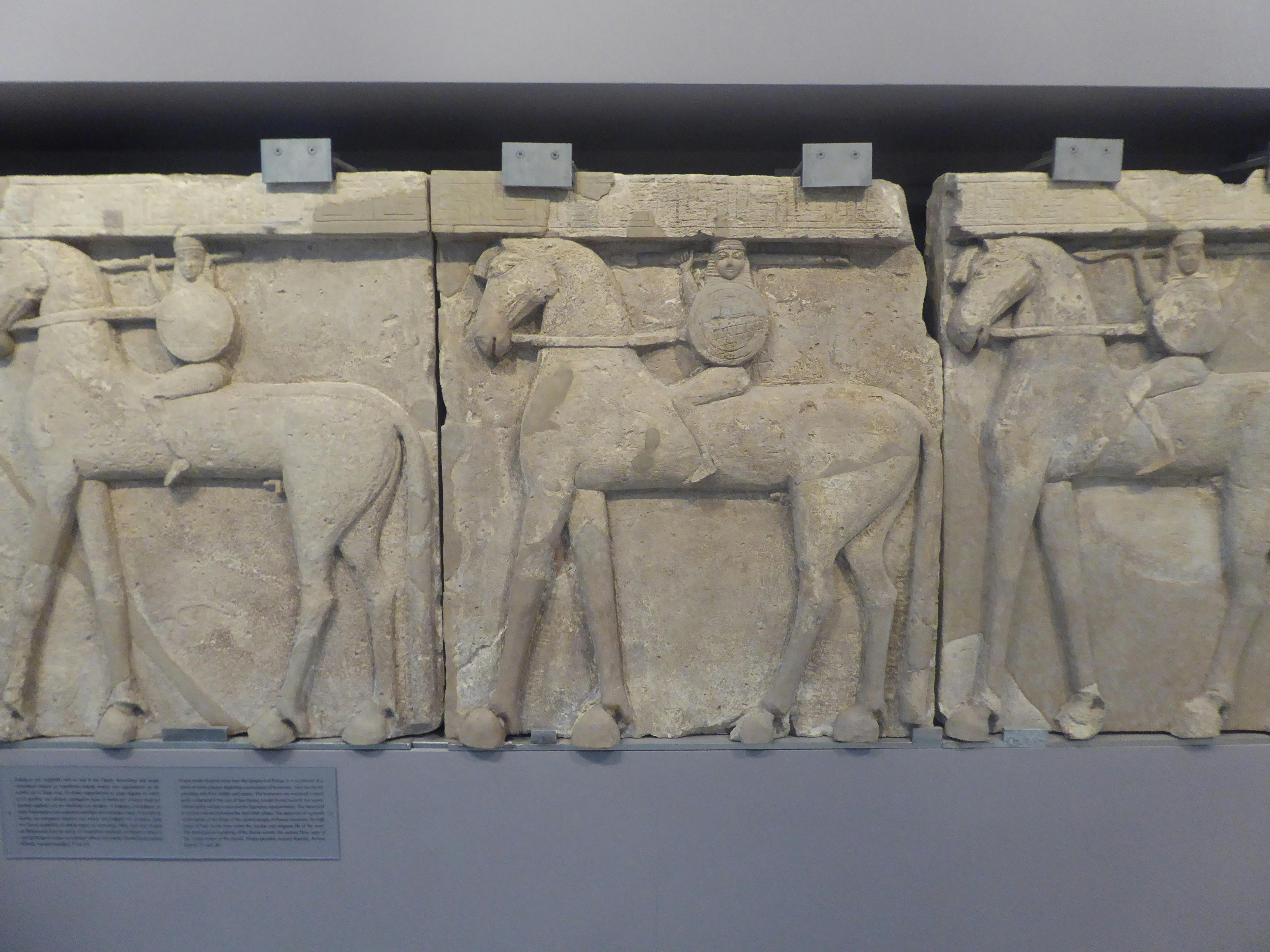 Trois anciens reliefs en pierre représentant des cavaliers sur des chevaux, montés sur le mur d'un musée. Plaque de texte visible en dessous.