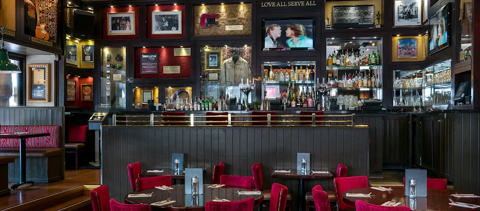 A bar interior with red chairs, tables set for dining, a well-stocked bar, framed memorabilia on the wall, and a TV showing a movie scene.