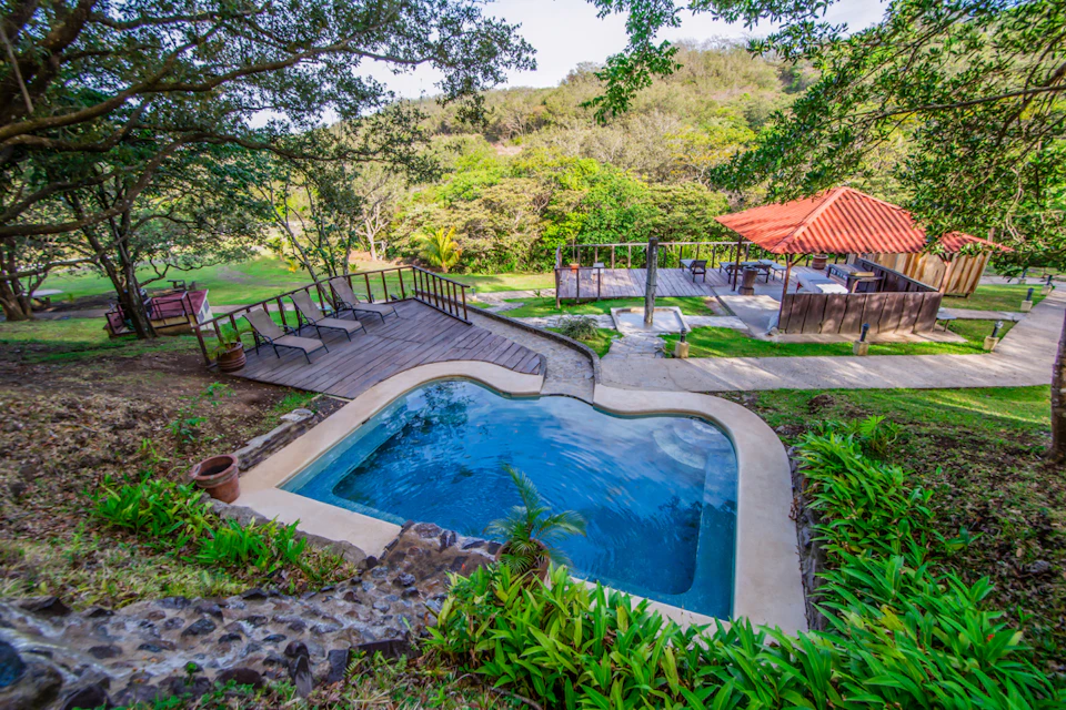 Enclosed pool surrounded by greenery, wooden deck with lounge chairs, and a gazebo with a red roof in a lush, forested area.