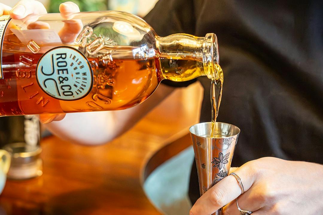 A person pouring Roe & Co whiskey into a metal jigger, with a close-up view of the bottle and hands.