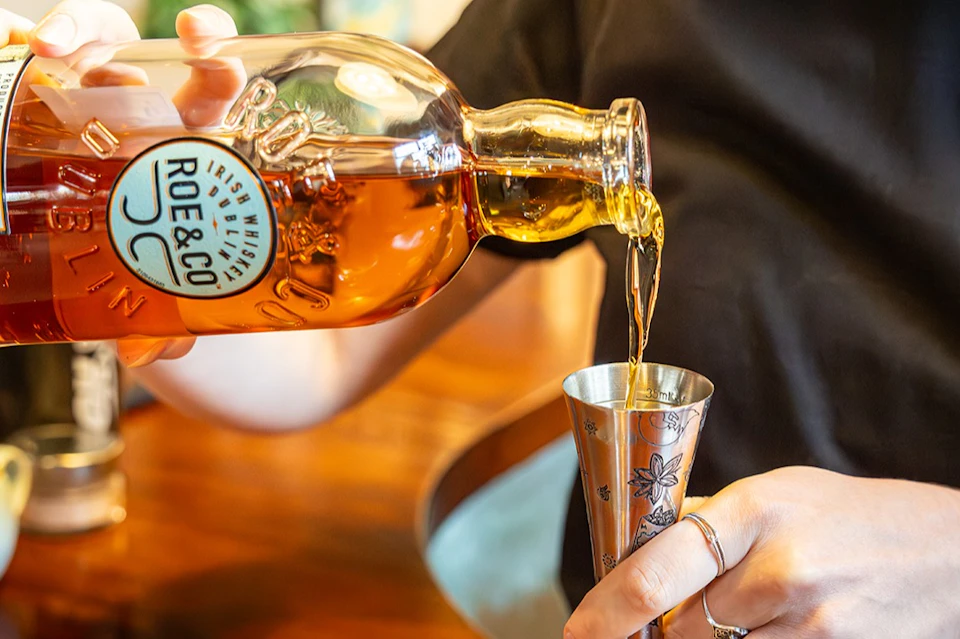 A person pouring Roe & Co whiskey into a metal jigger, with a close-up view of the bottle and hands.