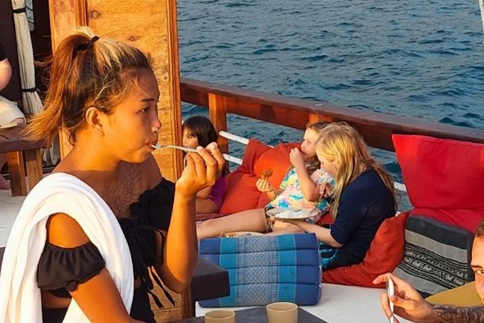 People enjoying food and drinks on a boat deck during sunset, with water and shoreline visible in the background.