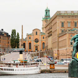 En scen vid vattnet med historiska byggnader, en kyrka, en hög obelisk, en båt som dockats och en bevingad staty i förgrunden.