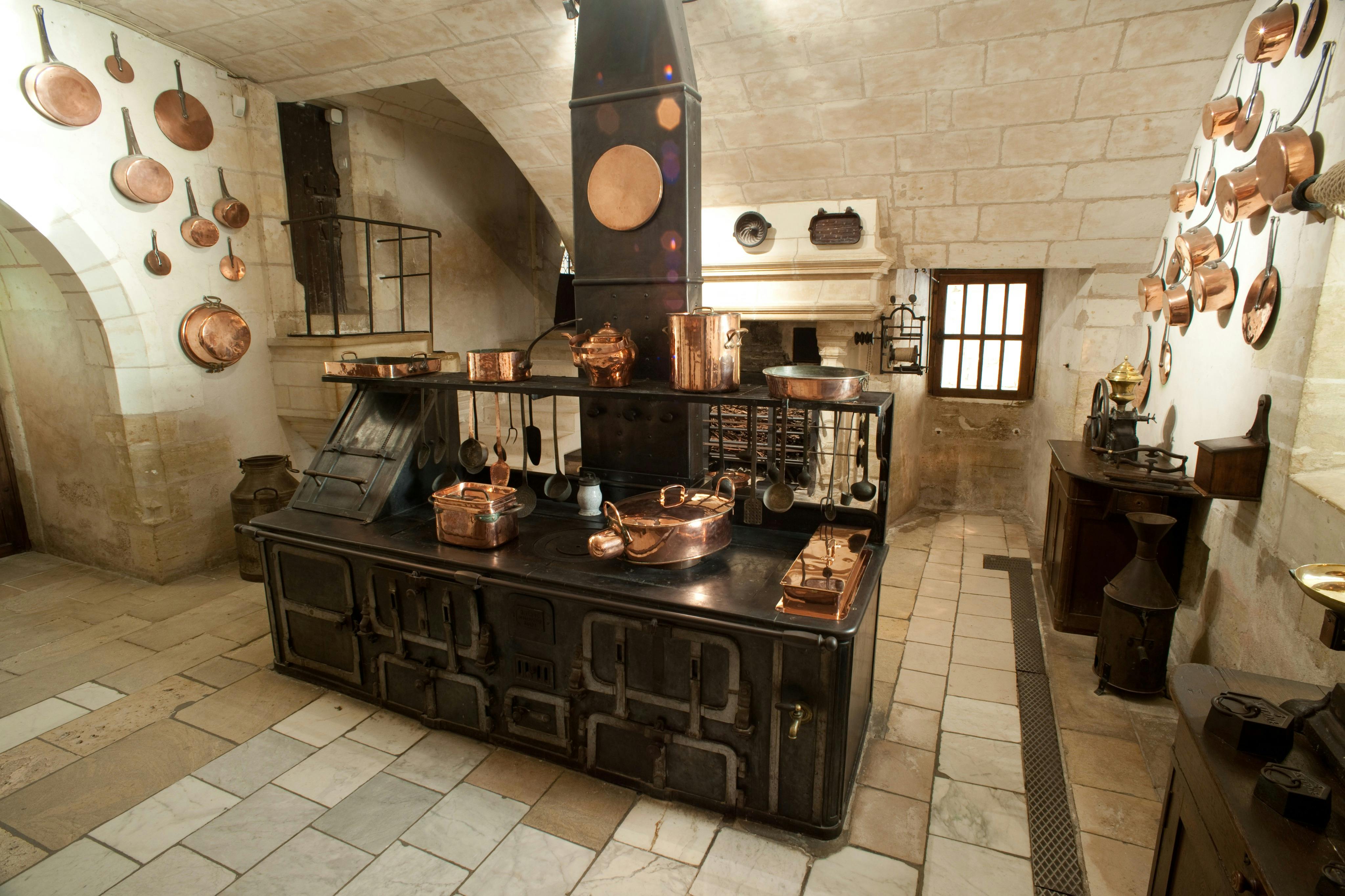 复古厨房，配有大型黑色炉灶、铜锅和平底锅、石墙和地板。 光线通过一扇小窗户进入。
