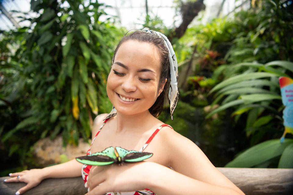 A woman smiling while a green and black butterfly rests on her hand in a lush greenhouse environment.