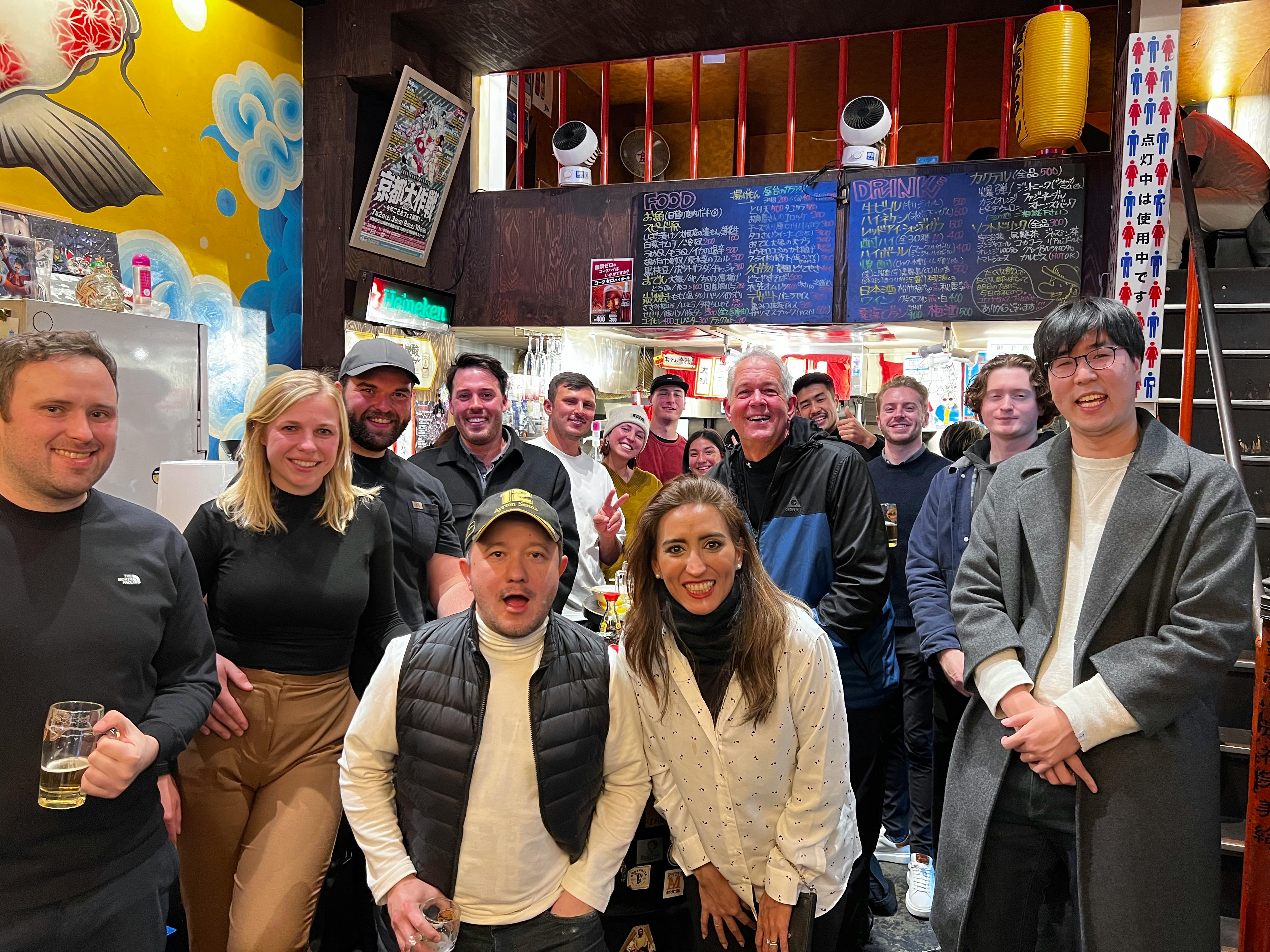 A group of people smiling and posing inside a lively restaurant with a colorful wall and a visible menu board.