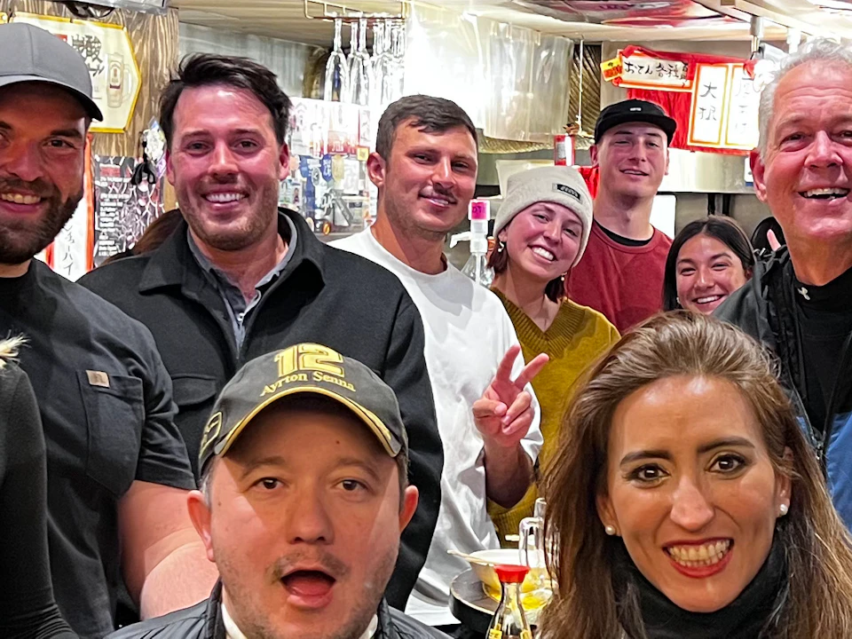 A group of people smiling and posing in a casual indoor setting, with colorful wall art and menu boards in the background.