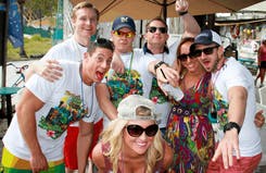 Un groupe de sept personnes, souriantes et posant énergiquement en plein air sous un auvent, portant des tenues colorées et des lunettes de soleil.