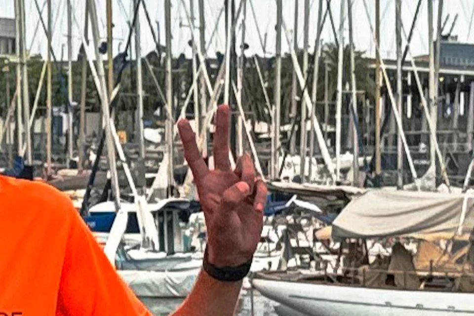Three people wearing helmets stand next to orange bicycles near a marina with boats and masts under a cloudy sky.