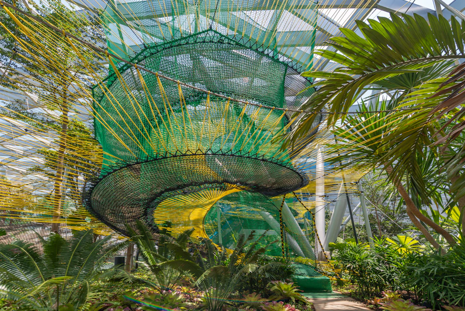A large, colorful netted structure with green, blue, and yellow hues sits among lush tropical plants inside a conservatory.
