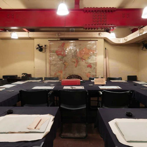 Stort konferensrum med U-format bord, svarta stolar och kartor. Papper och pennor ligger utspridda på bordet.