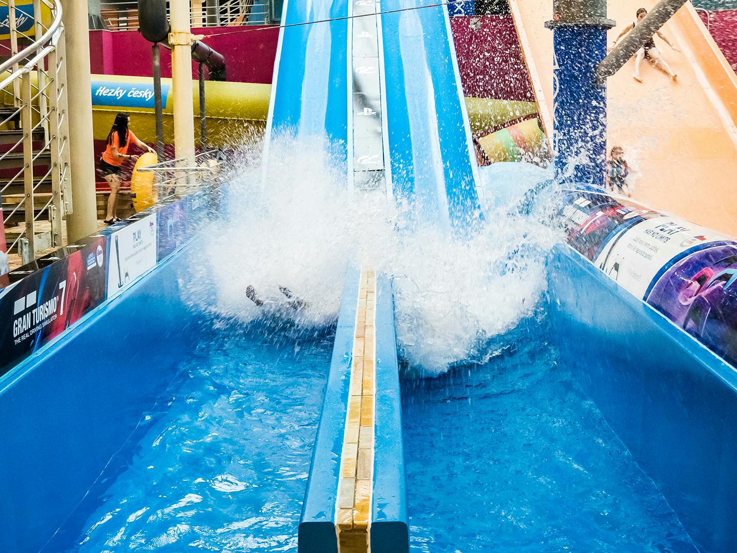Two people splash into the water at the bottom of a blue waterslide in an indoor recreational area.