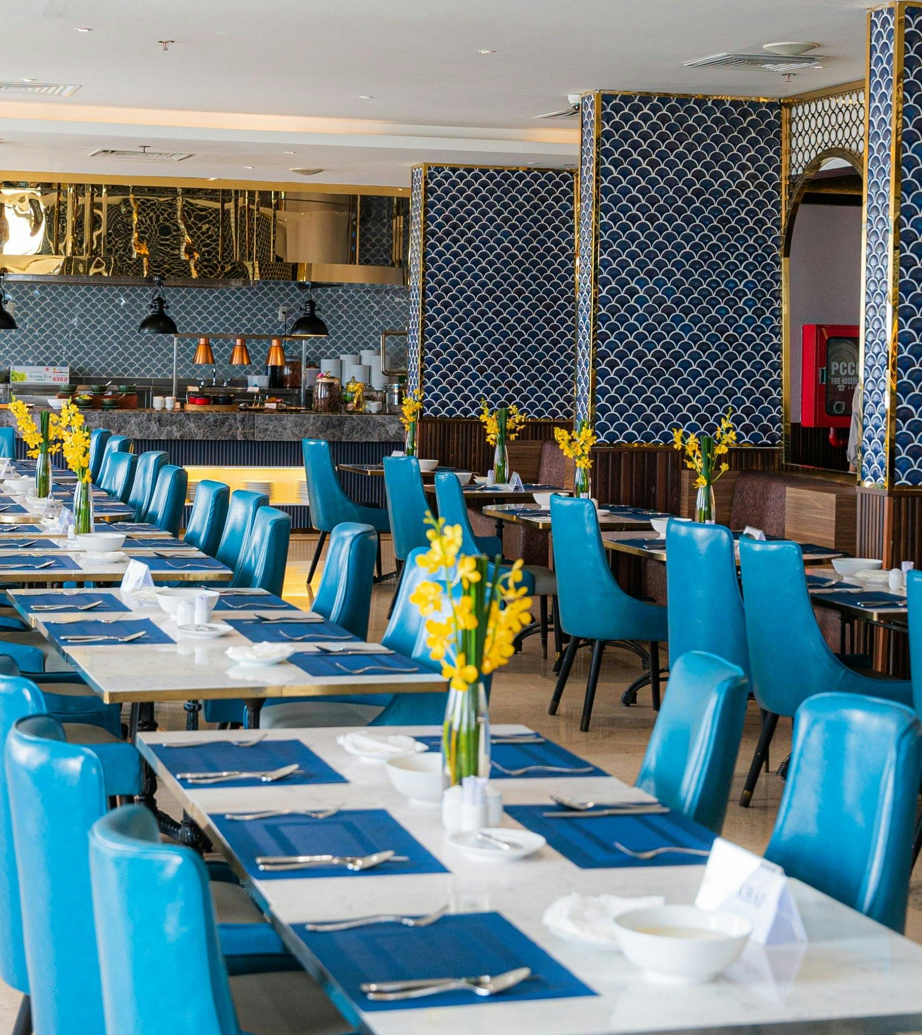 A restaurant interior with blue chairs, white tables set with tableware, and vases with yellow flowers as centerpieces.