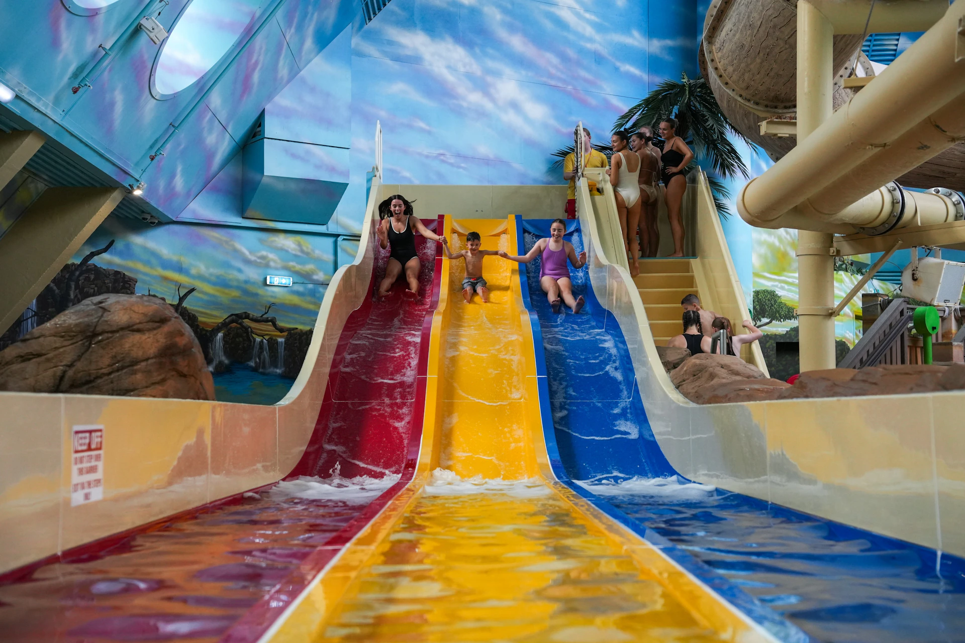 Children and adults sliding down colorful water slides in an indoor water park with themed decor and a painted sky backdrop.