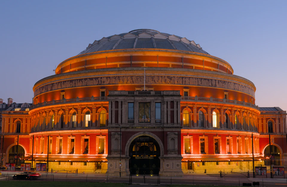 Large, illuminated circular building with ornate façade and dome-shaped roof at dusk. Arched entrance and detailed architectural features.