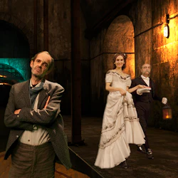 Tre personer i en svagt upplyst tunnel; en man med vikta armar i förgrunden och ett par klädda i formell klädsel i bakgrunden.