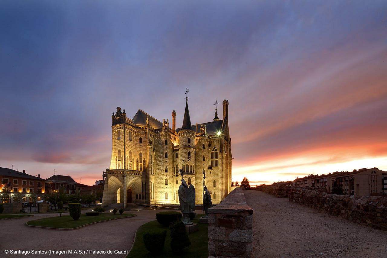 Un château illuminé sous un ciel crépusculaire avec une statue devant et un chemin de pierre menant à l'entrée.