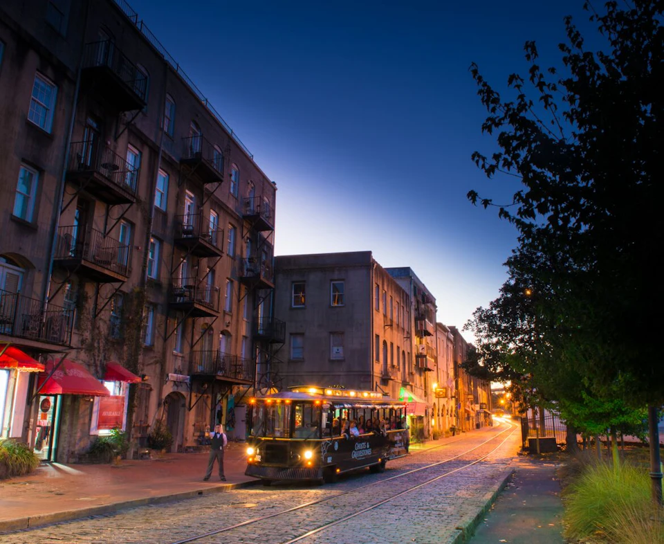 A streetcar moves down a cobblestone street at dusk, with old buildings and lit storefronts. Trees line the sidewalk.
