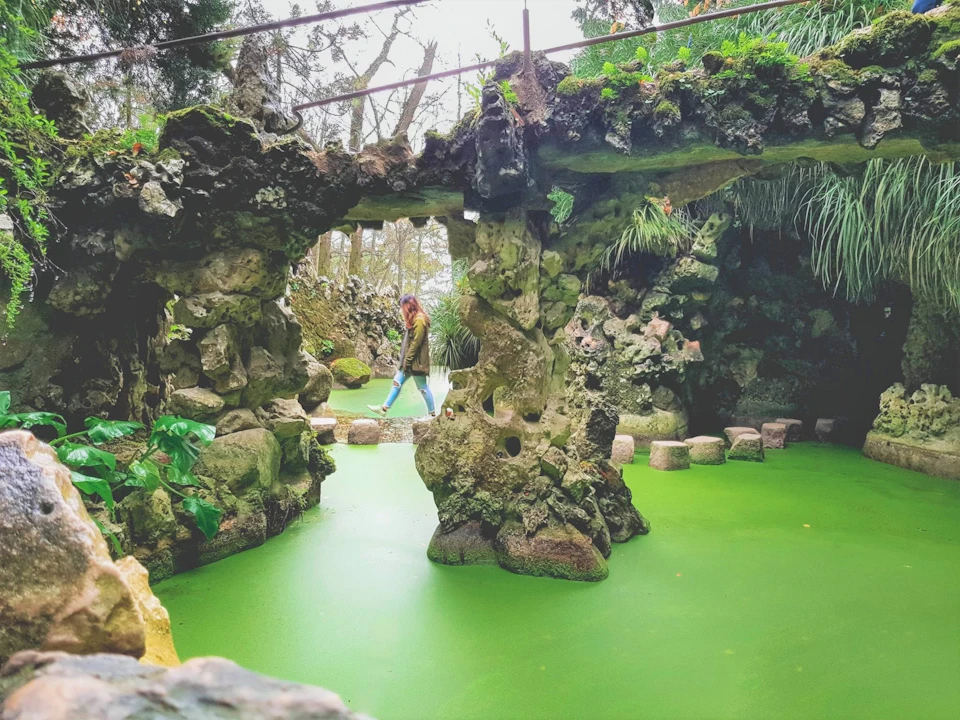 A person walks across stone steps above a green pond surrounded by rocky, moss-covered formations and lush greenery.