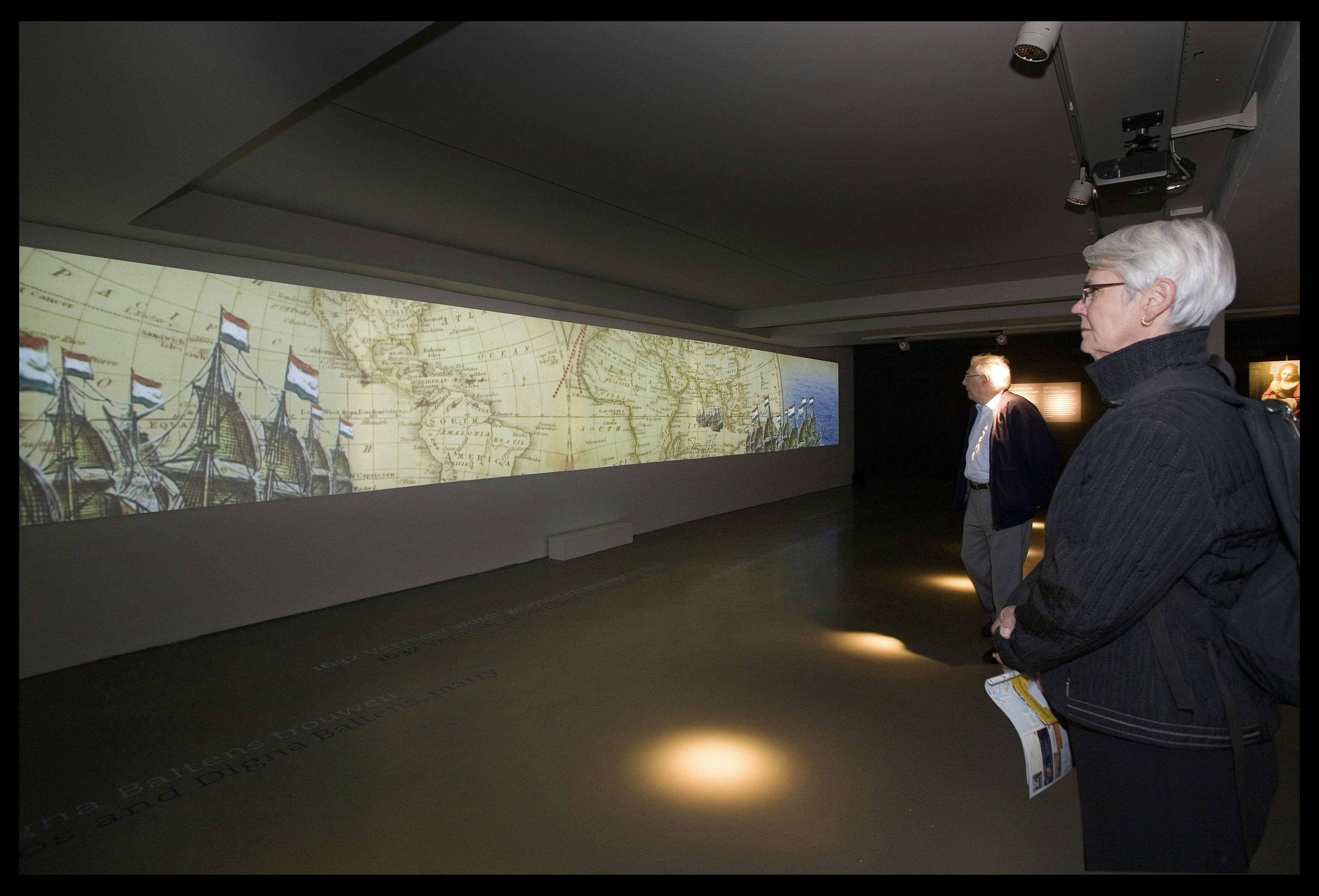 Duas pessoas numa sala de exposições pouco iluminada a olhar para um grande mapa histórico projetado na parede.