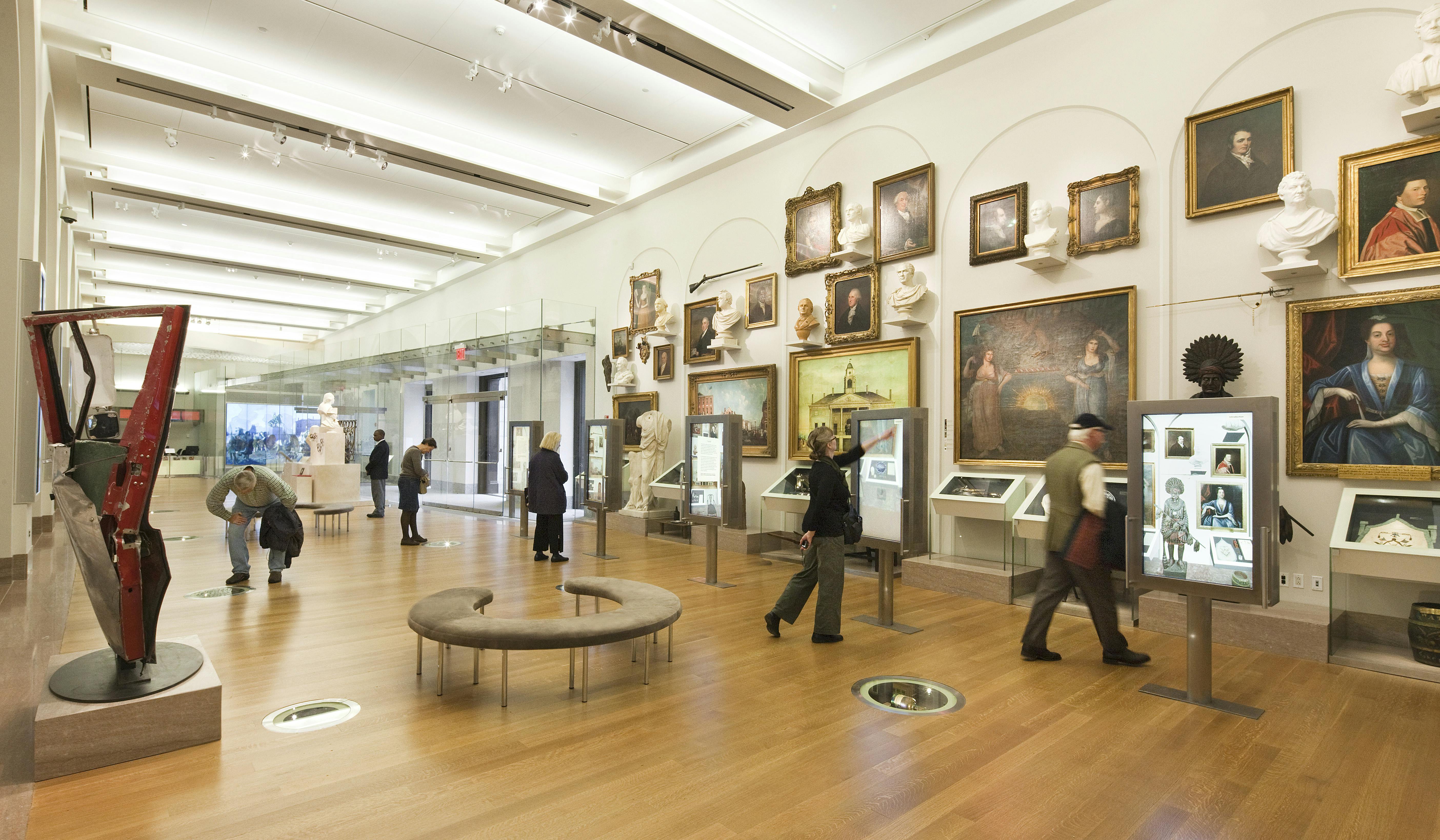 Άνθρωποι περπατούν σε μια γκαλερί τέχνης με τοιχογραφίες, γλυπτά και προθήκες σε ξύλινο πάτωμα.