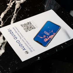 Ett papper med titeln "LJUDGUIDE" med en QR-kod och en smartphone som visar en bild ligger på en svart marmoryta.