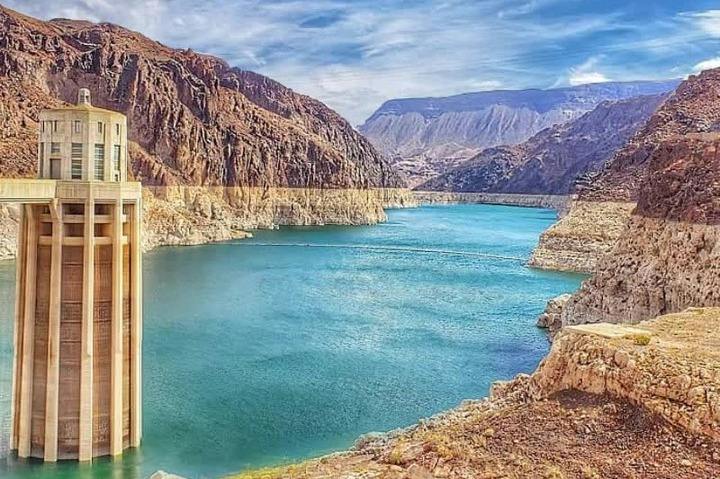 A scenic view of a dam with a reservoir surrounded by rocky cliffs and distant mountains under a partly cloudy sky.
