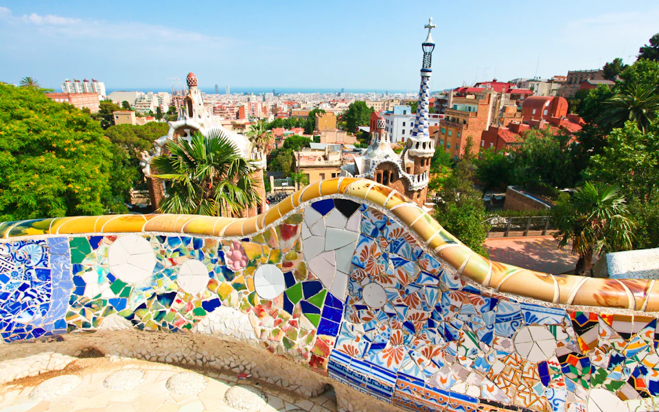 Colorful mosaic-tiled bench overlooking a cityscape with unique architecture, palm trees, and distant ocean.