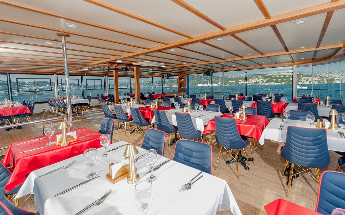 Elegant dining area with blue cushioned chairs, red and white tablecloths, set tables, and large windows overlooking the sea.