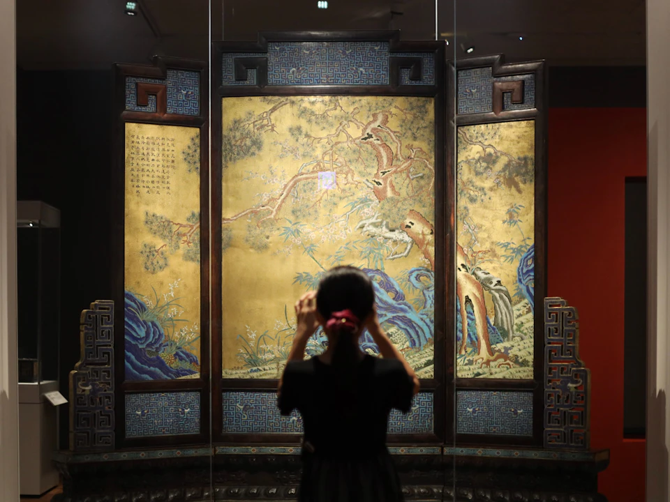 A person stands in front of an ornate screen with painted panels depicting animals and foliage, in a museum setting.