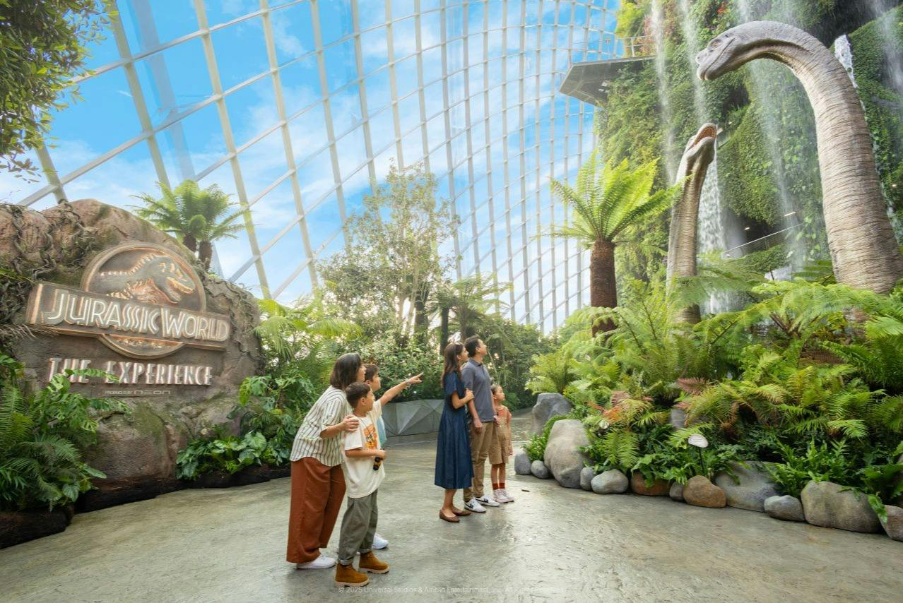 A family observes a dinosaur and waterfall in an indoor jungle-themed exhibit with a glass dome ceiling.