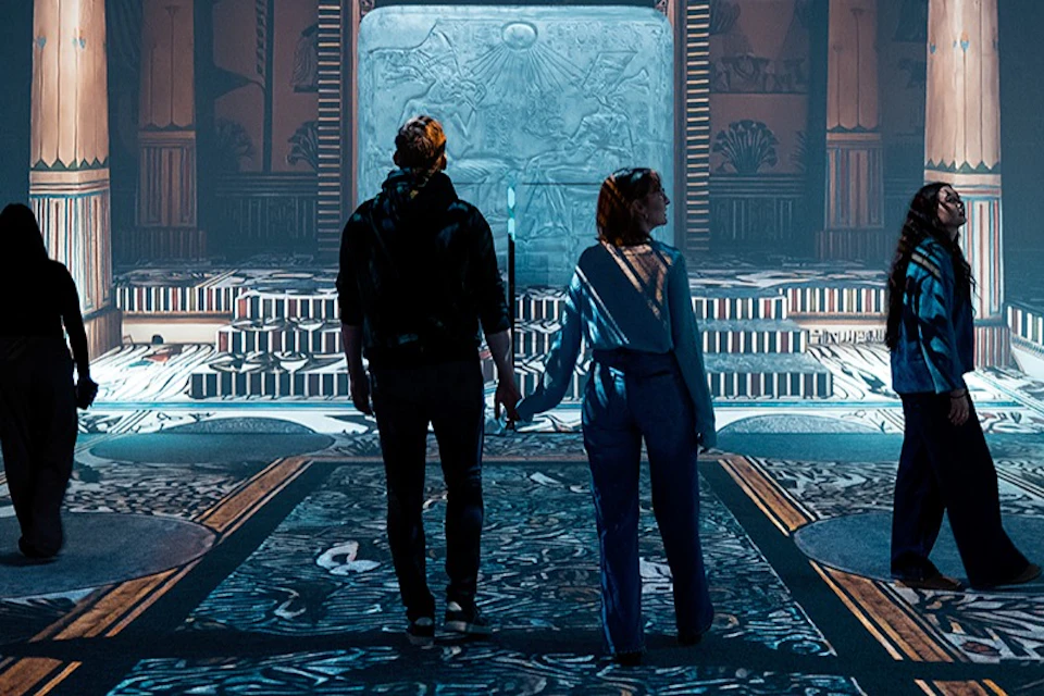 Two people stand hand in hand in a decorative hall with intricate patterns and a large illuminated relief in the background.