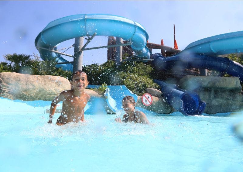 Two children splash in a pool at the base of a blue waterslide, with greenery and blue sky in the background.