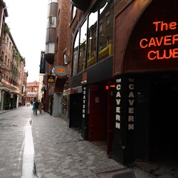 En smal gata med våta kullerstenar, flankerad av gamla tegelbyggnader, med "The Cavern Club" och "Audio Club" neonskyltar.