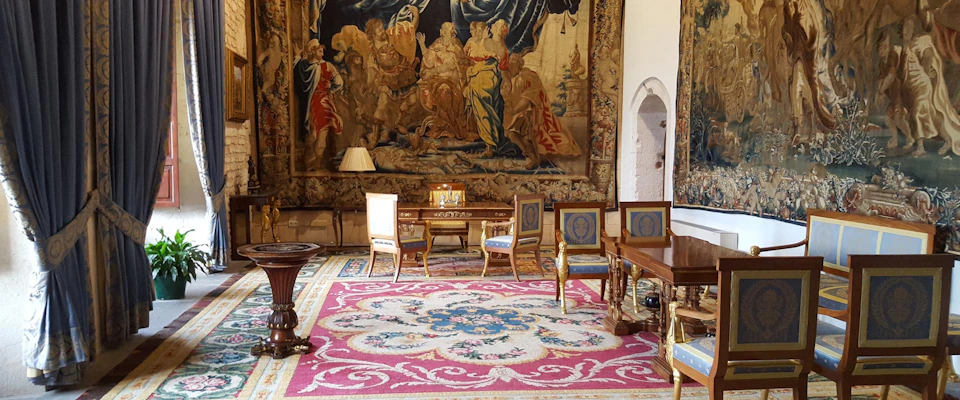 A room with ornate tapestries, decorative chairs, a wooden desk, a table lamp, and a richly patterned rug on the floor.