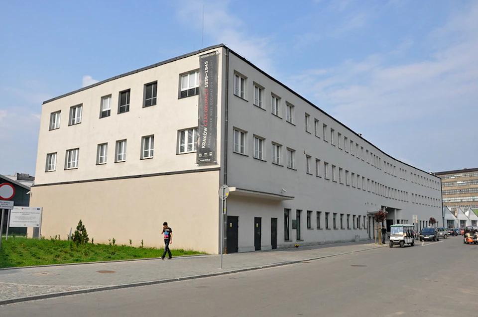 Rectangular, white building with many windows and a vertical banner. Person walking on the sidewalk. Clear blue sky.