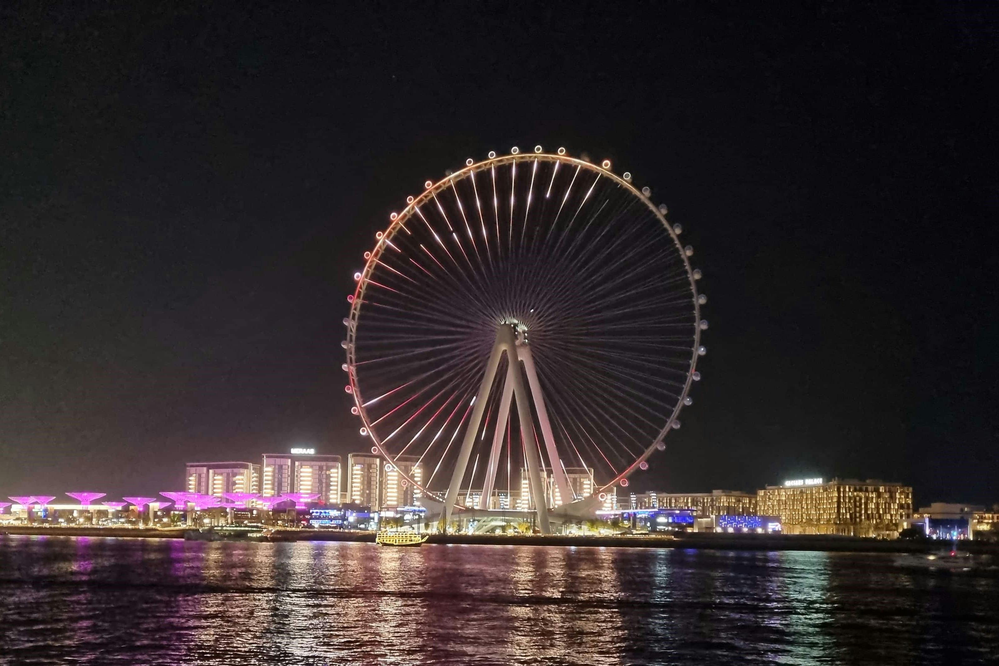 A large illuminated Ferris wheel at night with city lights and reflections on the water in the foreground.