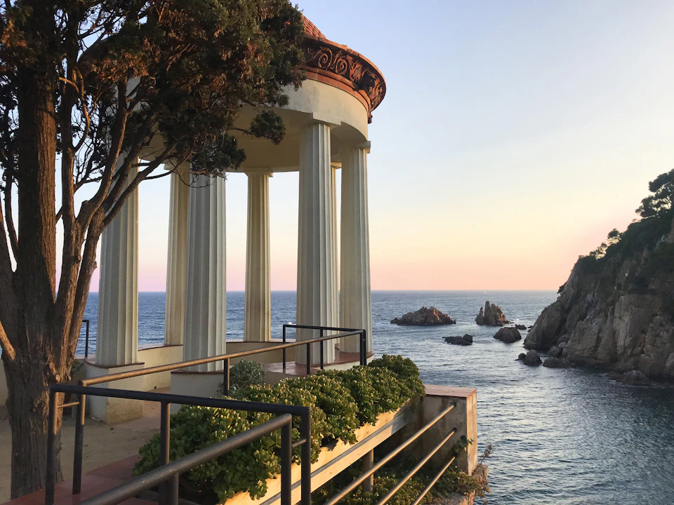 A columned pavilion overlooking the ocean at sunset, with rocky outcrops and lush greenery in the foreground.
