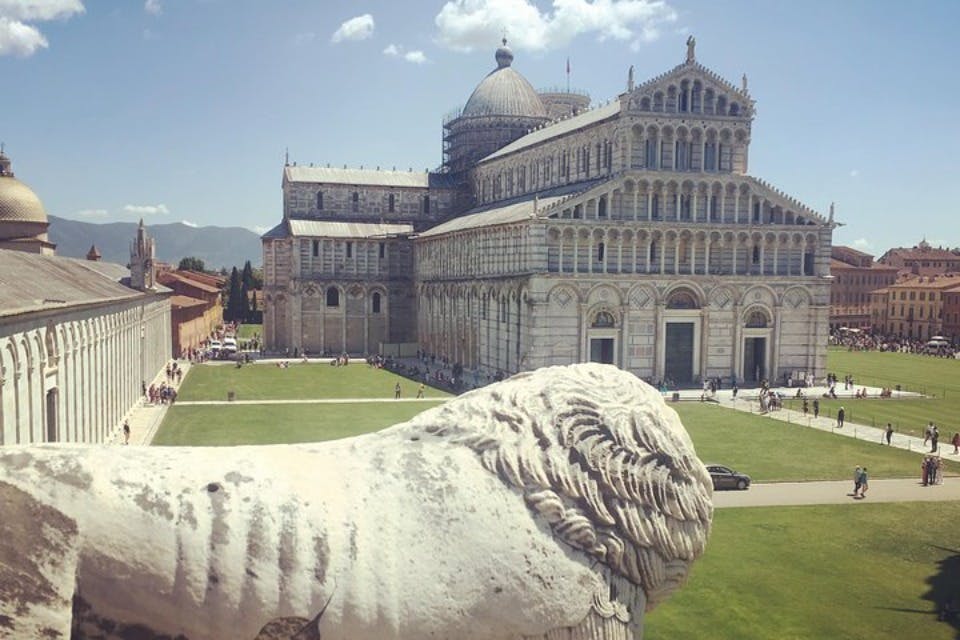 A view of the Pisa Cathedral with a large white stone sculpture in the foreground and people walking on the lawn.