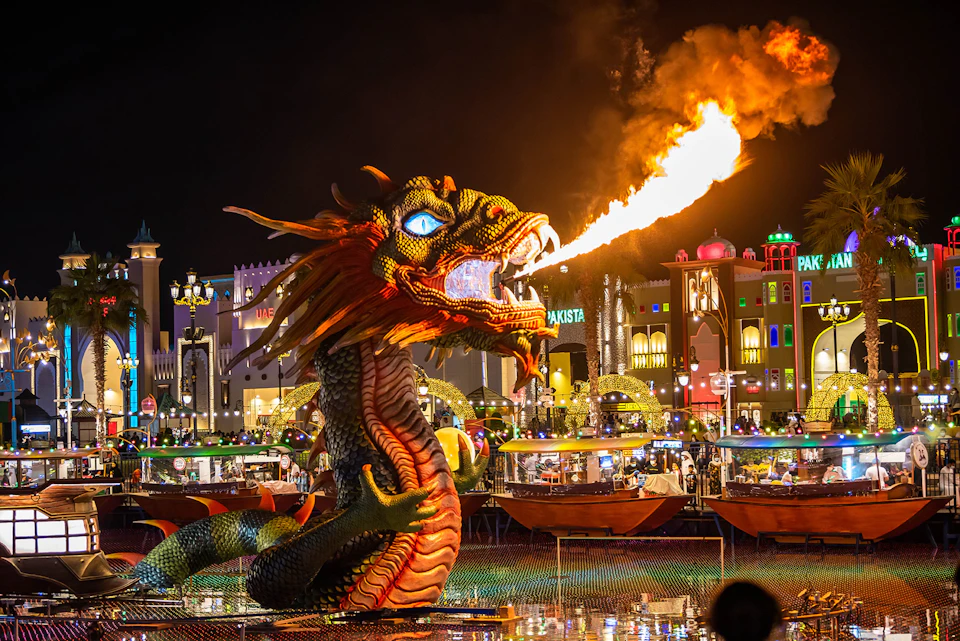 A large dragon sculpture breathes fire at night in a lively, colorful outdoor market with buildings and people in the background.