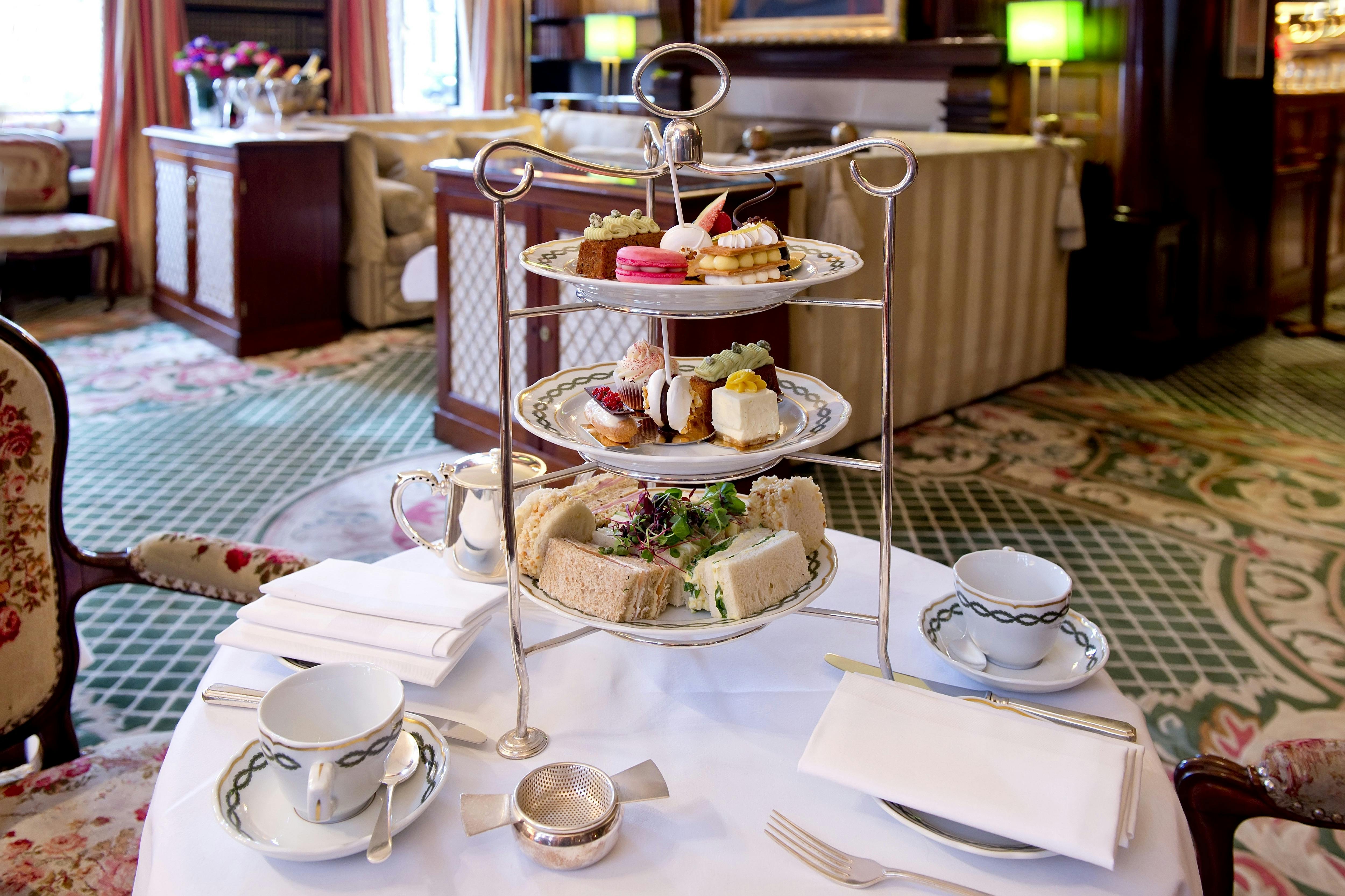 Elegant tea setup featuring a three-tier stand with sandwiches, pastries, and desserts; teacup and saucer on the table in a cozy room.