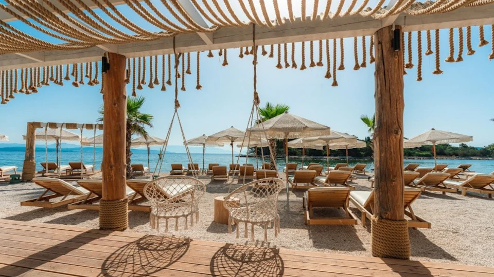 Beach scene with hanging wicker chairs, wooden loungers under straw parasols, and a view of the ocean.