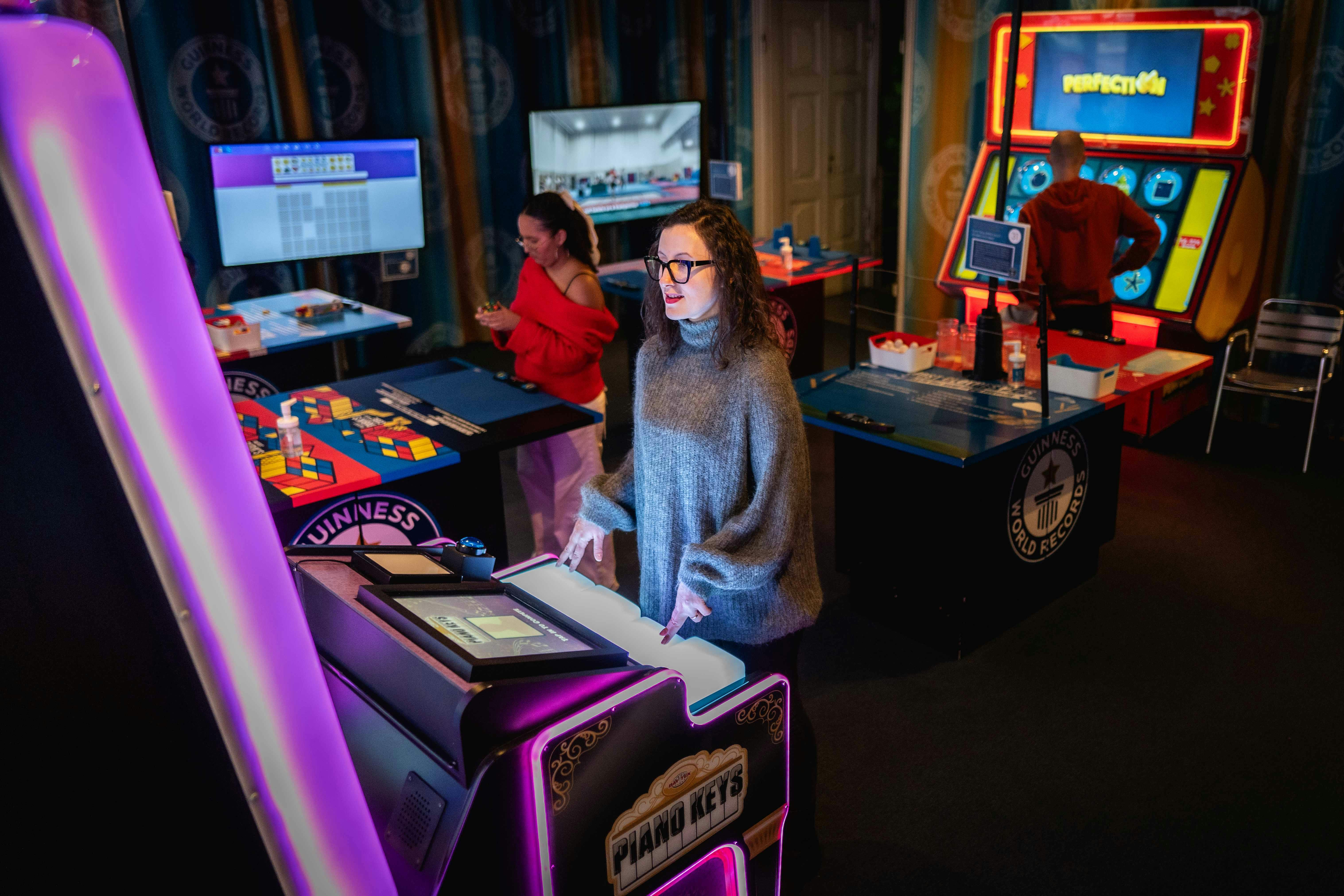 A woman plays a piano keys arcade game while another woman in a red sweater observes nearby tables with various games.