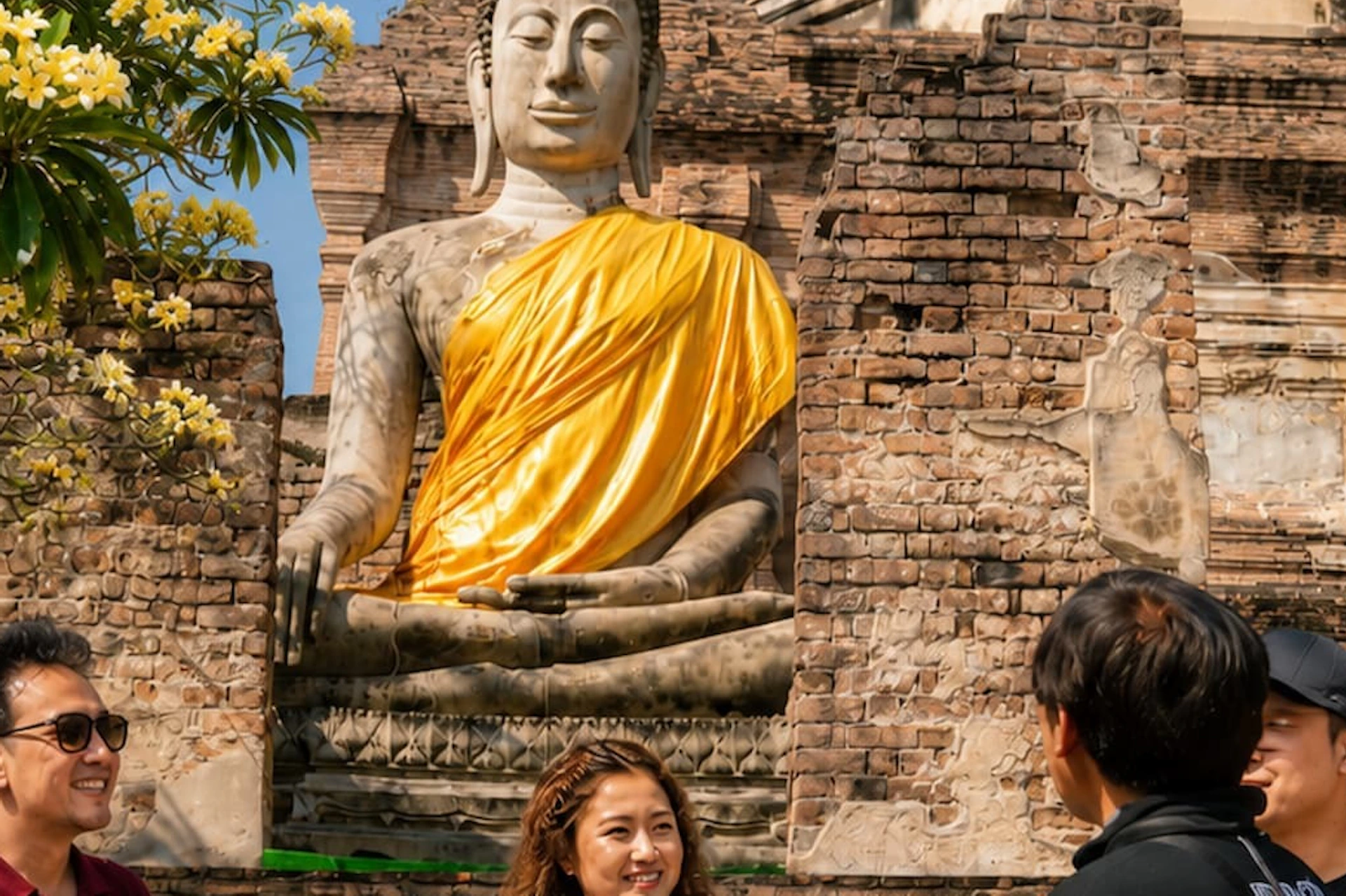 A group of people standing in front of a large seated Buddha statue draped in a yellow fabric, set against an ancient brick structure.