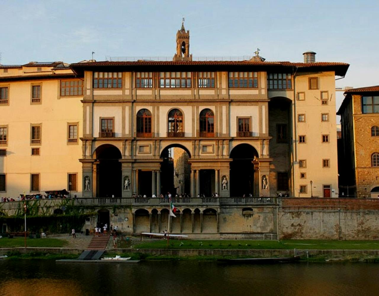 A grand riverside building with arched windows and statues, fronted by a grassy shoreline and a few people near the water.