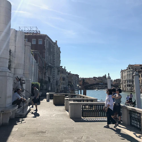 Människor går och vilar vid en kanal i Venedig med båtar och historiska byggnader i bakgrunden under en klar, blå himmel.