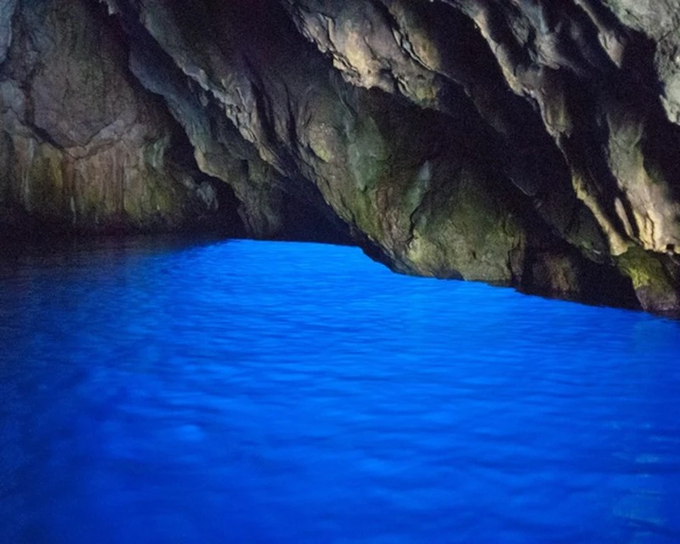 A cave with rocky walls and a vibrant blue, illuminated body of water inside.