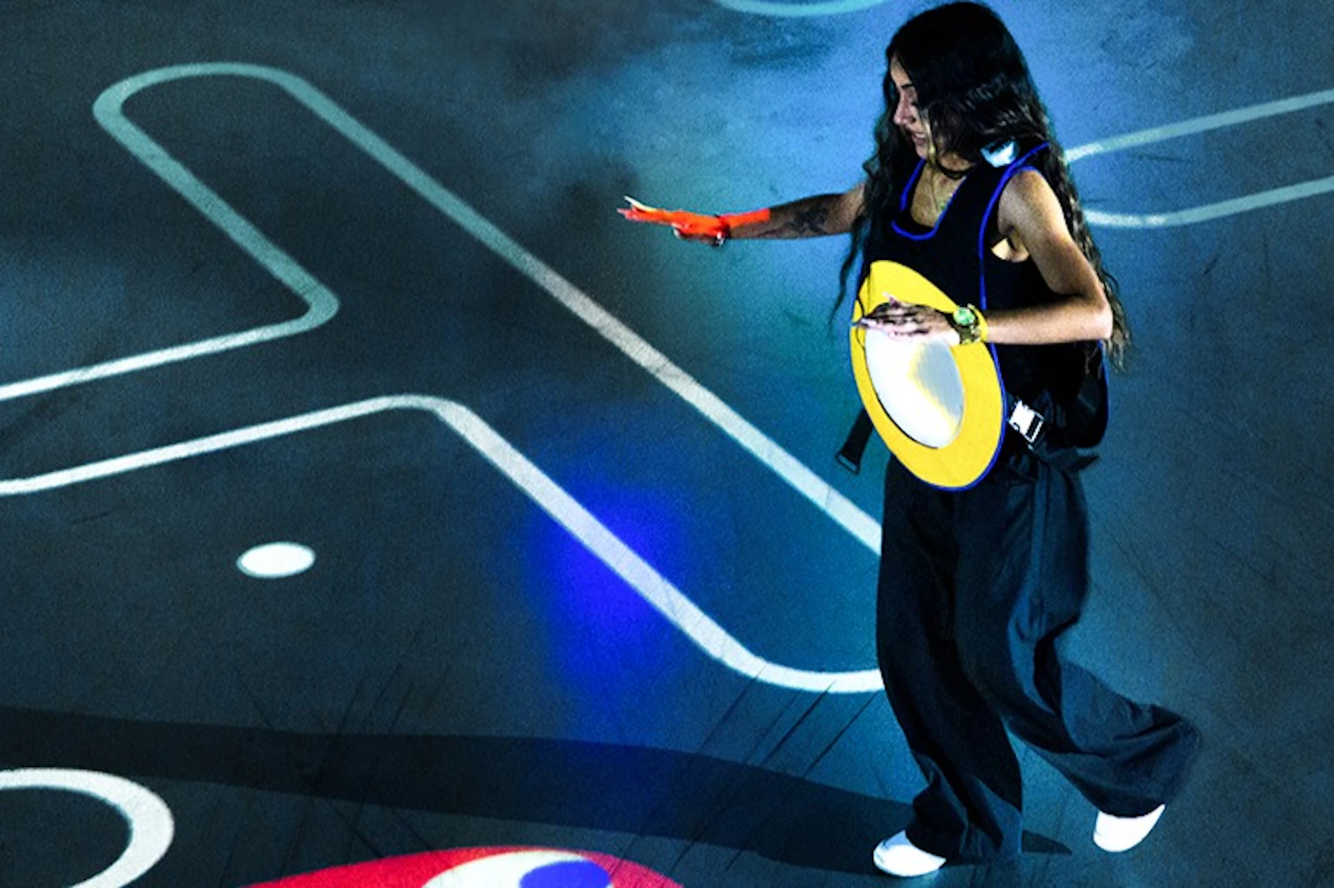 A woman with long hair walks on a street at night, holding a yellow circular object with one hand extended.