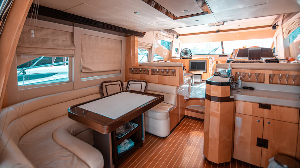 Luxurious boat interior with wooden flooring, a dining table with bench seating, a helm station, and large windows with blinds.